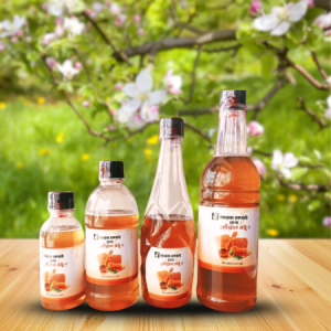 A display of four plastic bottles of natural honey labeled ""জায়ে রকমারি মধু" in varying sizes, placed on a wooden surface with a vibrant green garden background. Blooming white and pink apple blossoms on a tree branch are visible above, emphasizing the natural and organic setting.